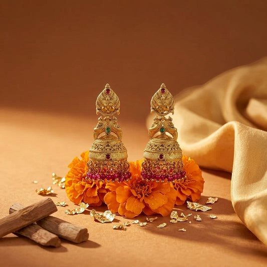 Lakx Fashion gold-tone brass jhumka earrings with deity motifs, green and ruby stones, and pink bead fringe on marigolds.