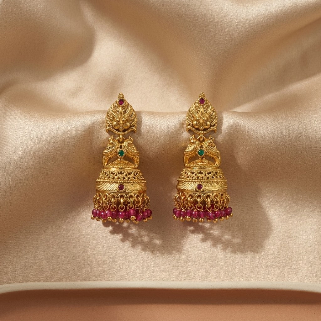 Lakx Fashion gold-tone brass jhumka earrings with deity motifs, green and ruby stones, and pink bead fringe on silk.