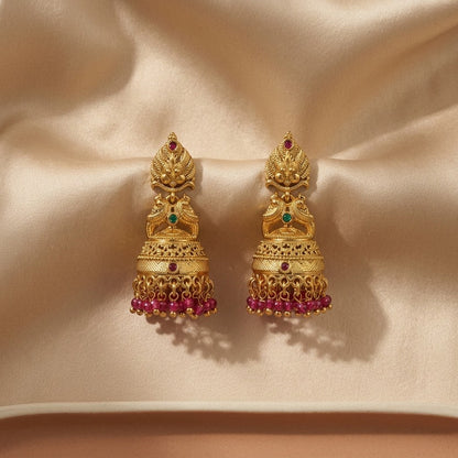 Lakx Fashion gold-tone brass jhumka earrings with deity motifs, green and ruby stones, and pink bead fringe on silk.