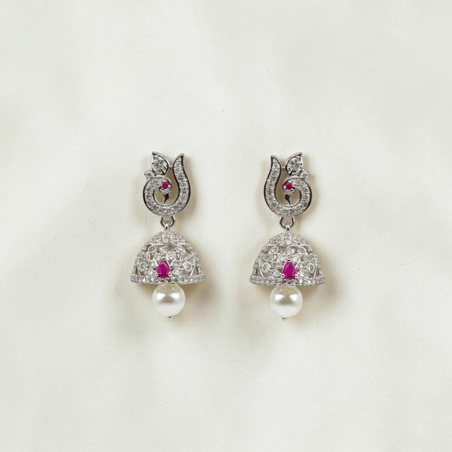 Lakx Fashion rhodium-tone jhumka earrings with ruby CZ stones, peacock motif, and pearl drops on a textured surface.