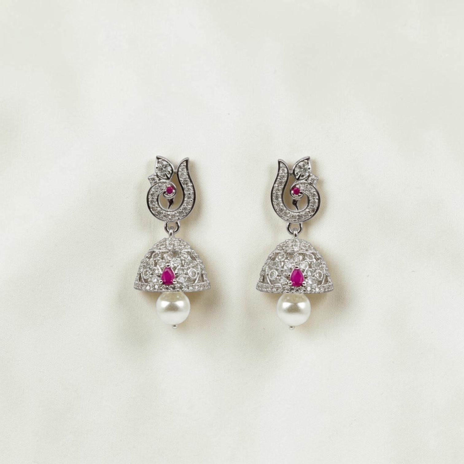 Lakx Fashion rhodium-tone jhumka earrings with ruby CZ stones, peacock motif, and pearl drops on a textured surface.