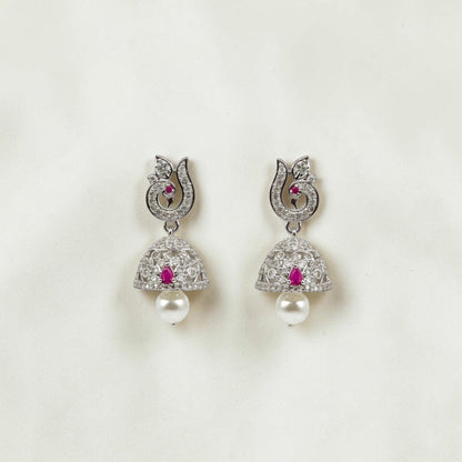 Lakx Fashion rhodium-tone jhumka earrings with ruby CZ stones, peacock motif, and pearl drops on a textured surface.