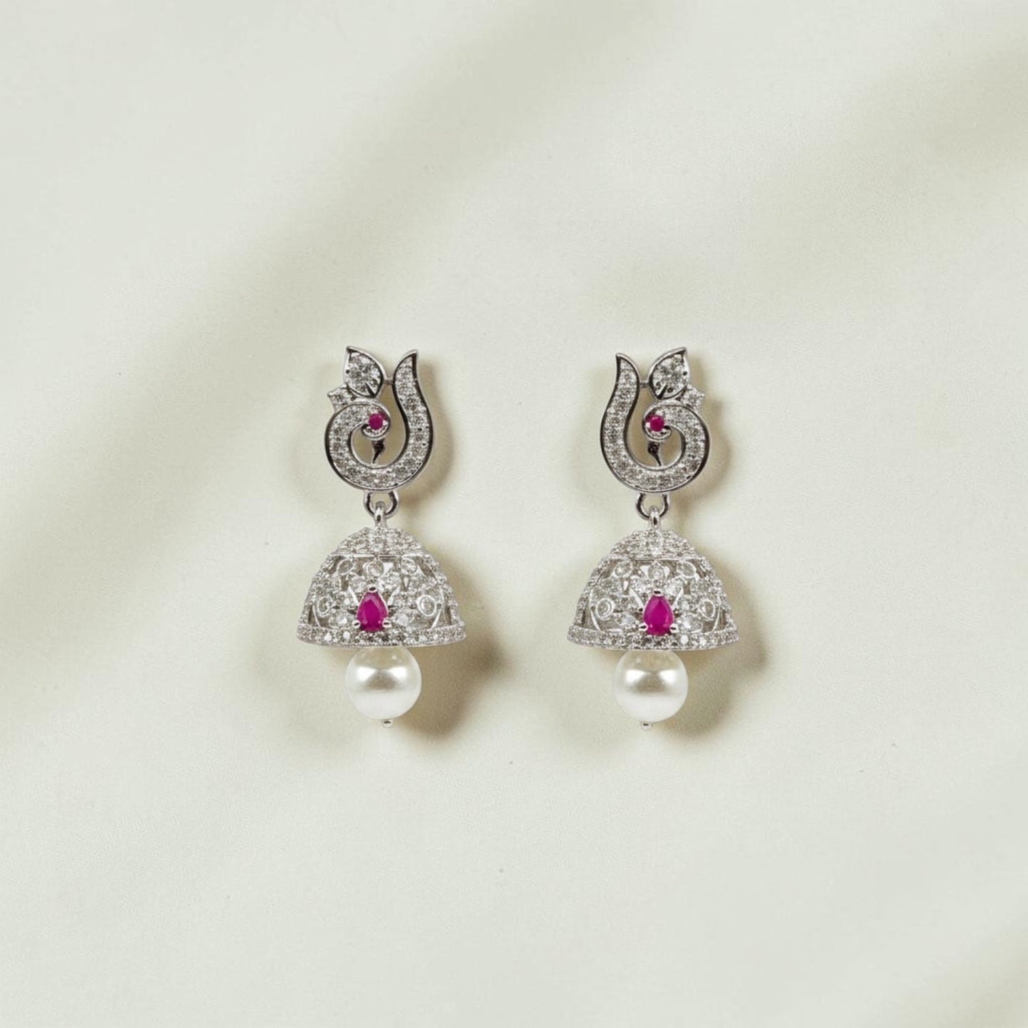 Lakx Fashion rhodium-tone brass jhumka earrings with ruby-colored stones, white CZ, and pearl drops on wood background.