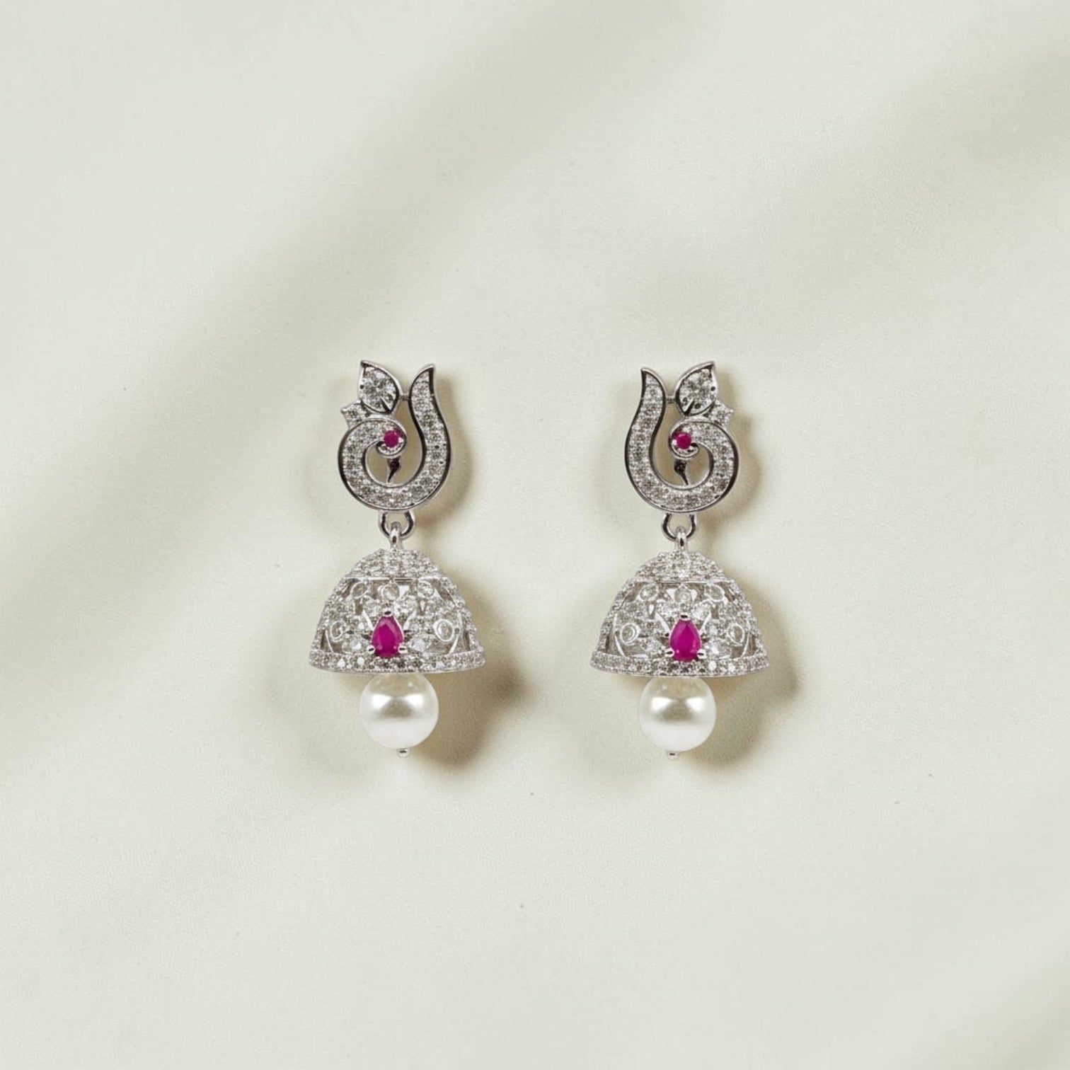 Lakx Fashion rhodium-tone brass jhumka earrings with ruby-colored stones, white CZ, and pearl drops on wood background.